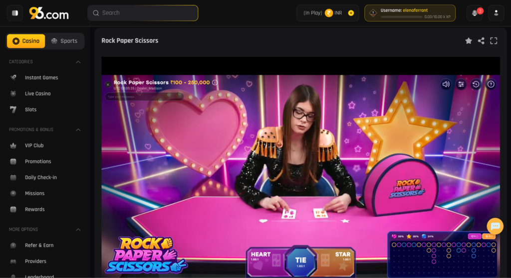 image - Woman in a sequined outfit sits at a pink game table, dealing cards against a neon pink stage with a large heart prop and star lights for Rock Paper Scissors.